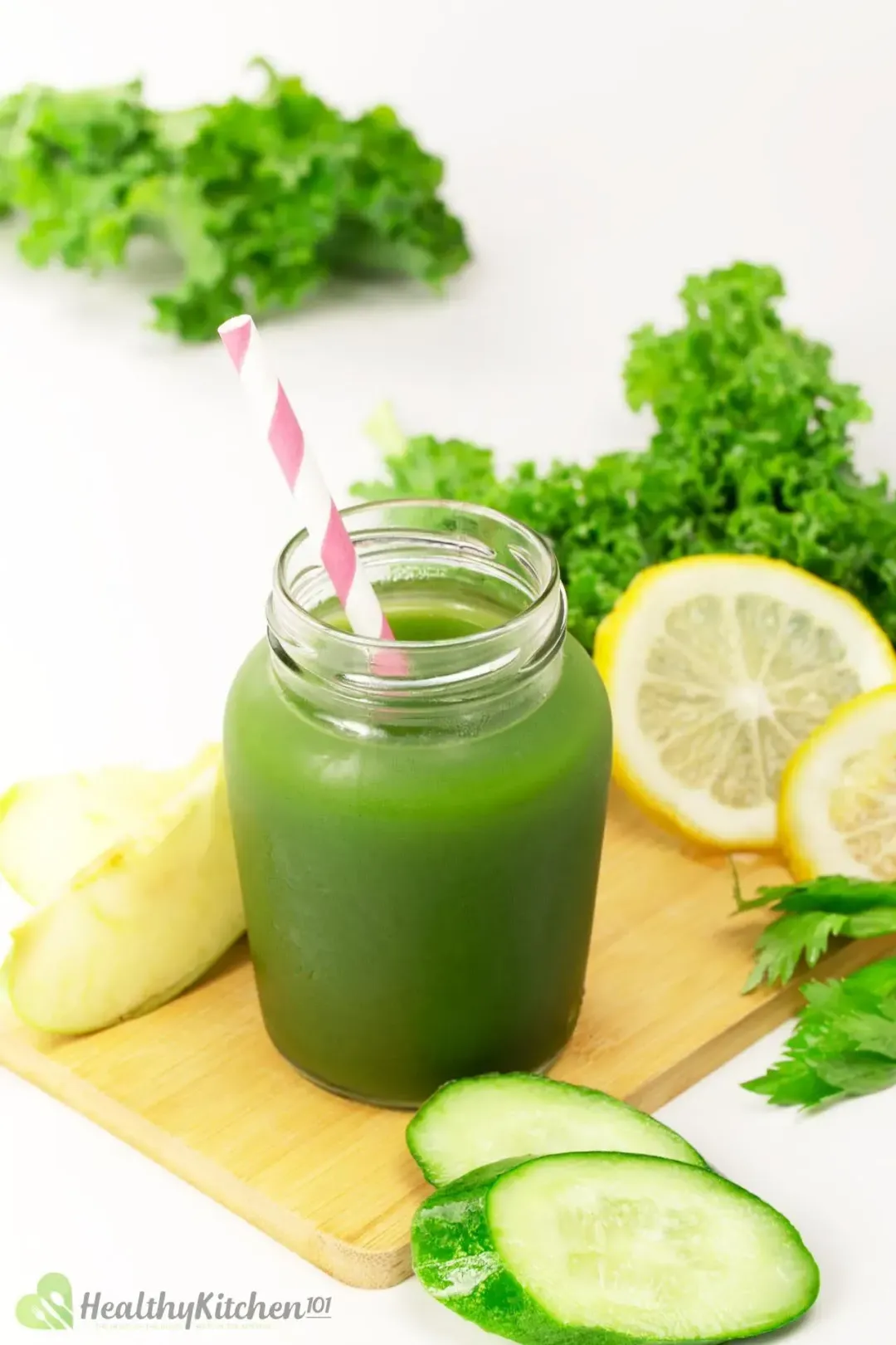 A cool jar of green vegetable juice with a pink-striped straw placed on a wooden board with cucumber slices, a peeled ginger knob, lemon slices, and kale leaves