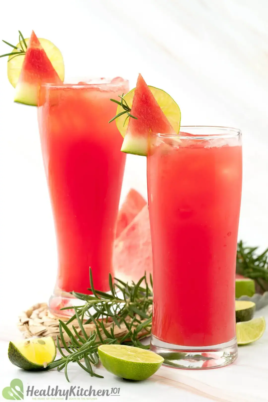 Two iced watermelon juice glasses held side by side, with watermelon wedges, limes, and rosemary as garnish