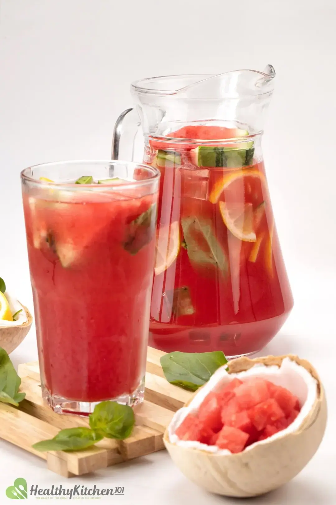 A jug and a glass of watermelon juice placed on a wooden board next to a coconut shell with watermelon cubes
