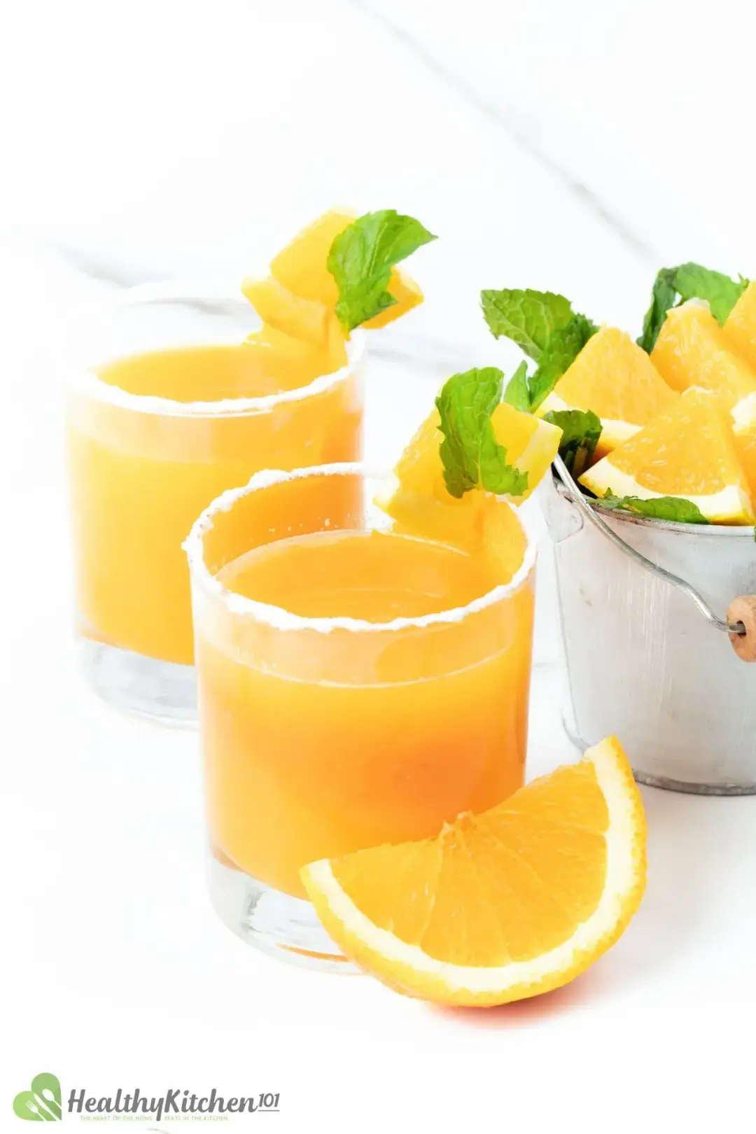 Two short glasses of whiskey and orange juice cocktail with mint sprigs and orange wedges around