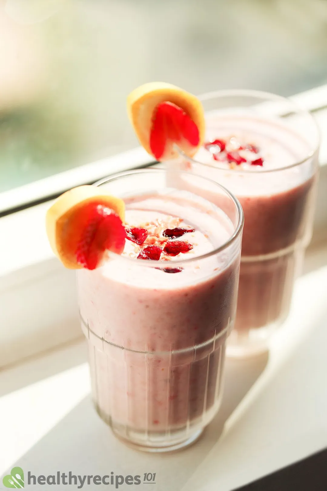 Two glasses of pink smoothie garnished with banana slices and strawberry slices laid near a window