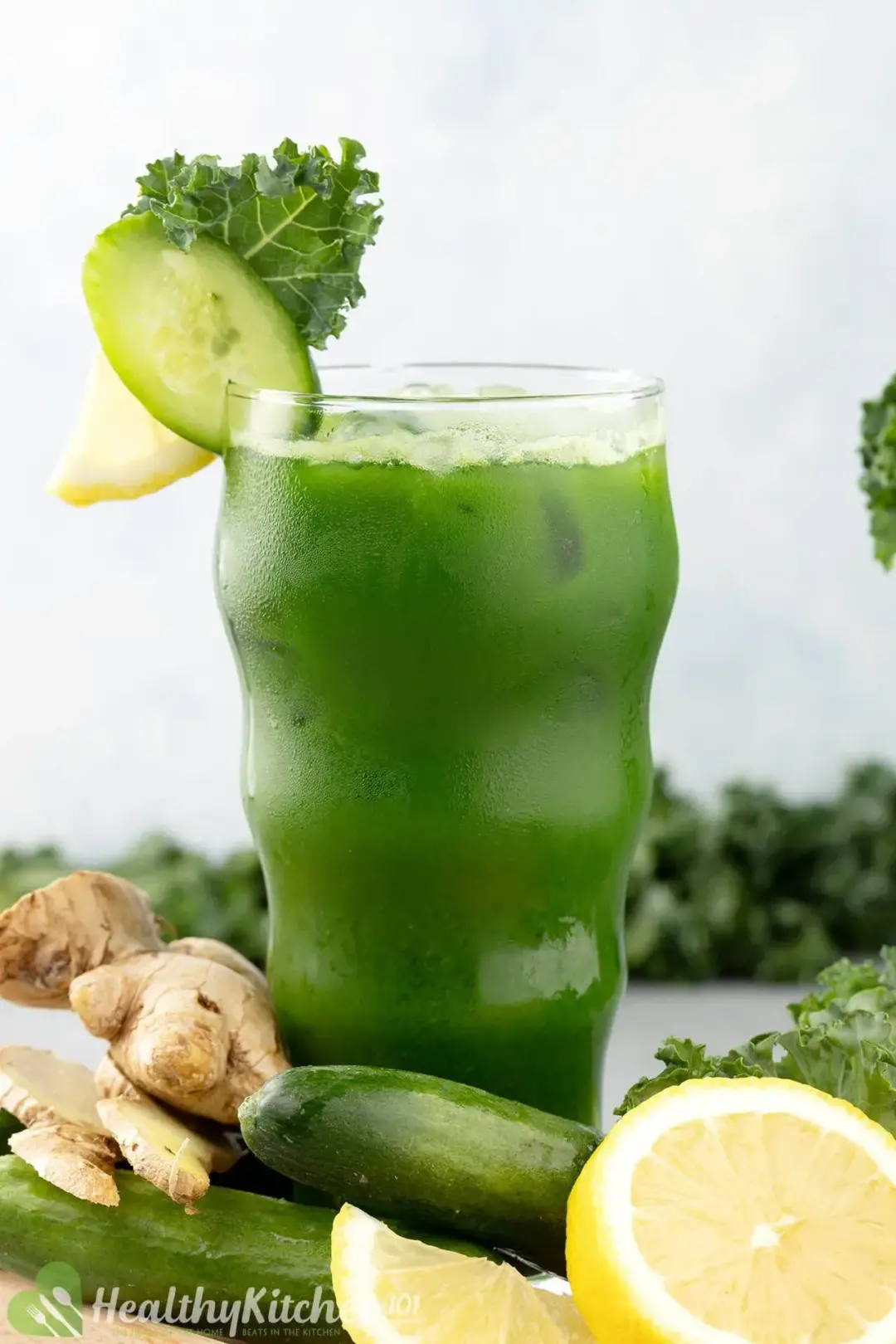 An iced glass of kale juice, garnished with cucumber slice, lemons, whole cucumbers, and a ginger knob