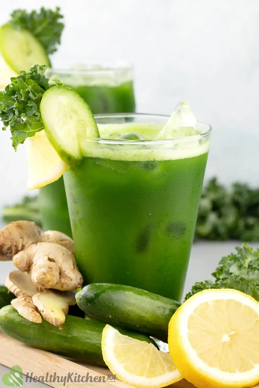 A close-up shot of a green juice glass with ice, next to some lemon wheels, cucumbers, and ginger