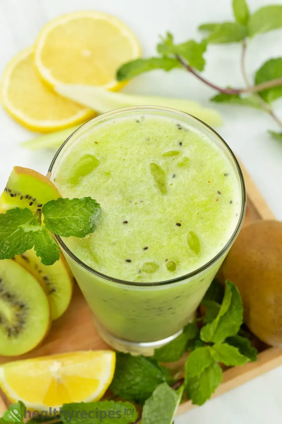 A glass of foamy kiwi juice on a wooden tray with lots of mints, kiwi slices, and and lemon wedge