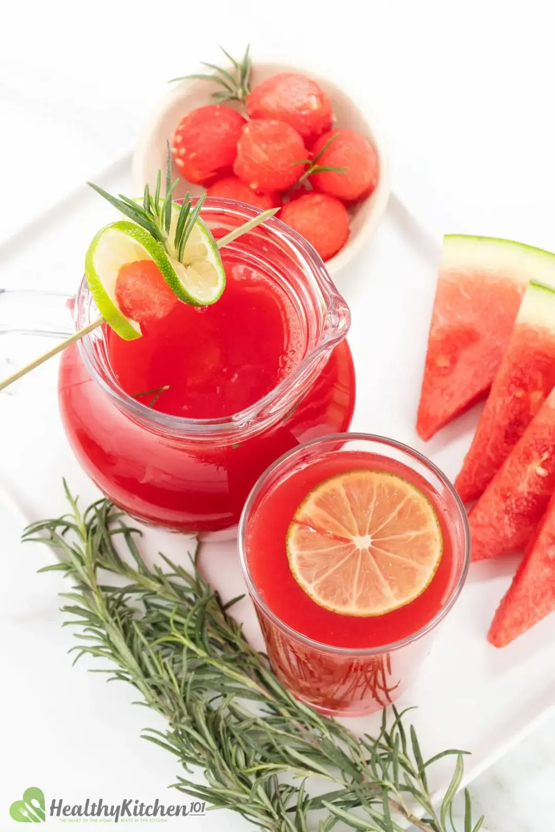 A white tray of watermelon juice pitcher, watermelon drink, watermelon balls, watermelon wedges, and rosemary stalks