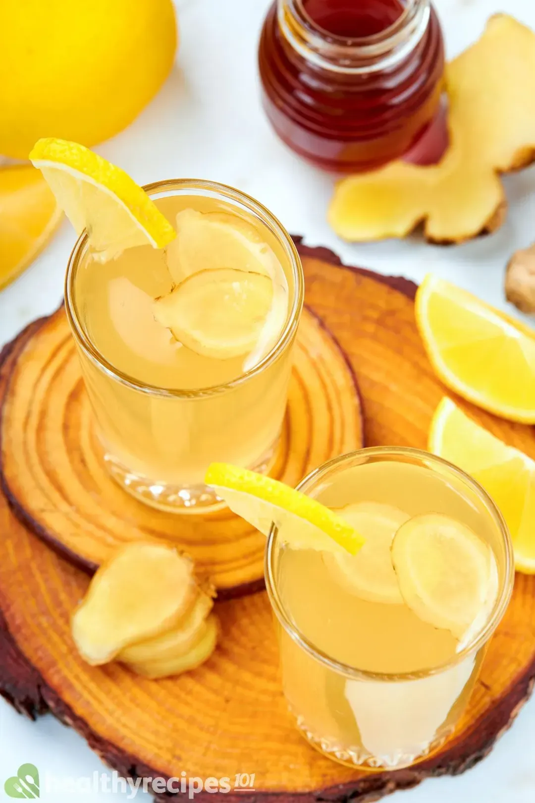 Two glasses of honey ginger vinegar garnished with ginger slices, lemon wedges, and next to a small honey jar