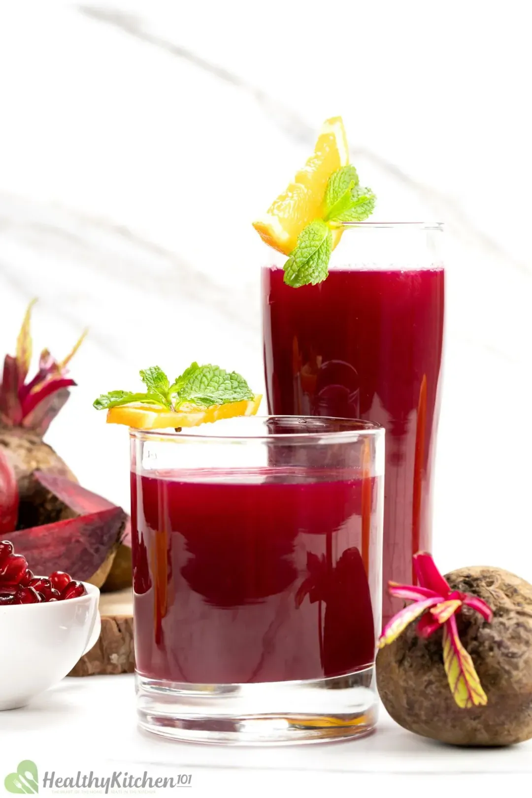 A short glass and a tall glass of beetroot juice, garnished with orange wedges, mints, and whole beetroots 