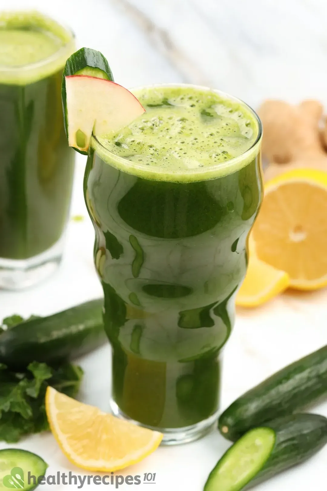 A glass of green juice in the center, surrounded by lemons, cucumbers, and other items in the background