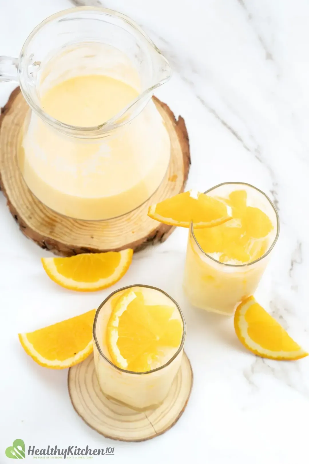 Two glasses of orange and milk drink next to a large pitcher and lots of orange wedges