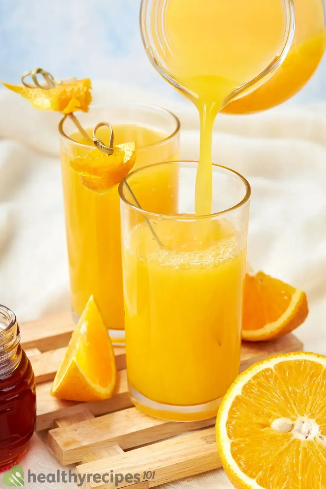 A pitcher pouring orange juice into a glass that’s next to an orange drink and lots of oranges