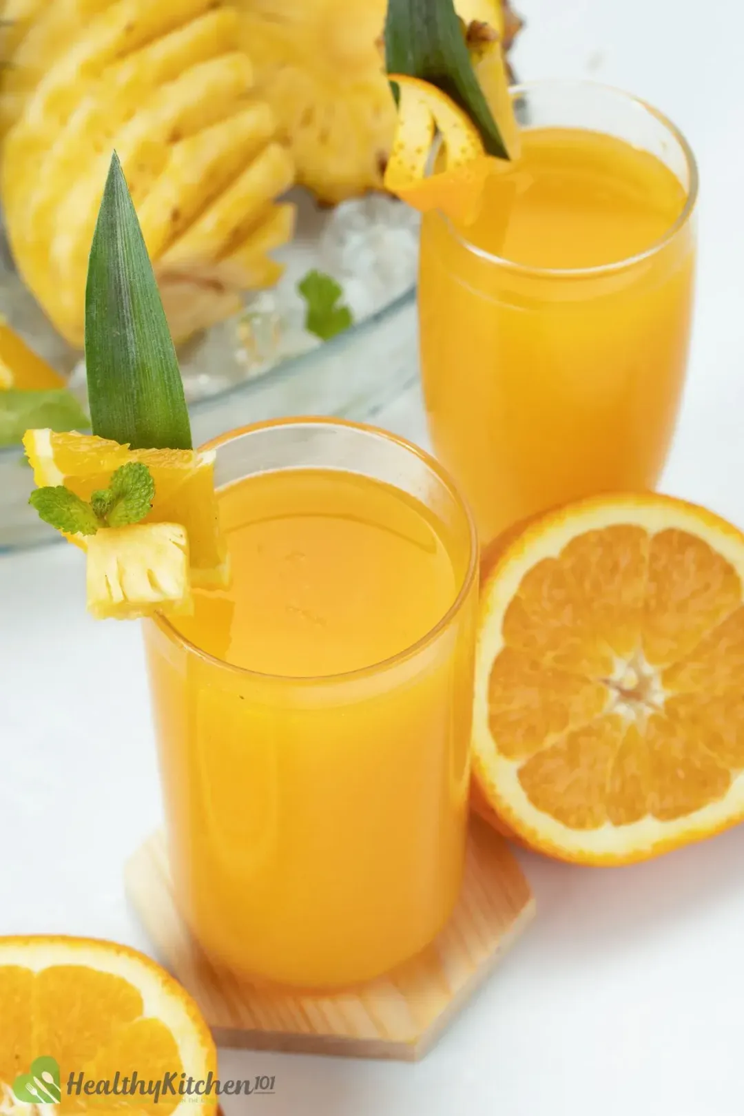 Two glasses of orange pineapple juice, next to half an orange and some peeled pineapples