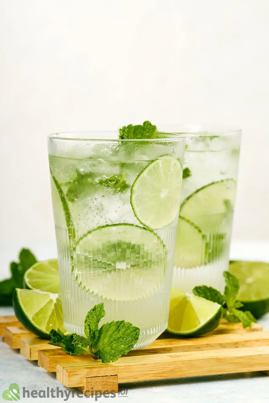 Two mojitos glasses of lime wheels, ice, mints, and bubbling soda, on a wooden tray with mints and limes