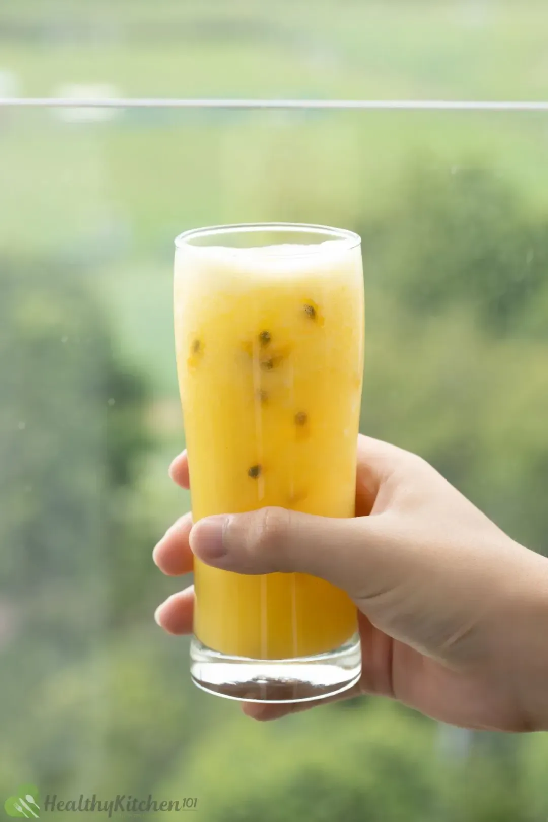 A hand holding a glass of passion fruit blended with mango by a window