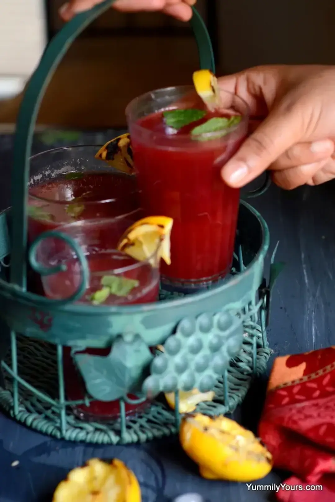 A hand picking up a glass of plum juice from a round drink tray, surrounded by pieces of lemons