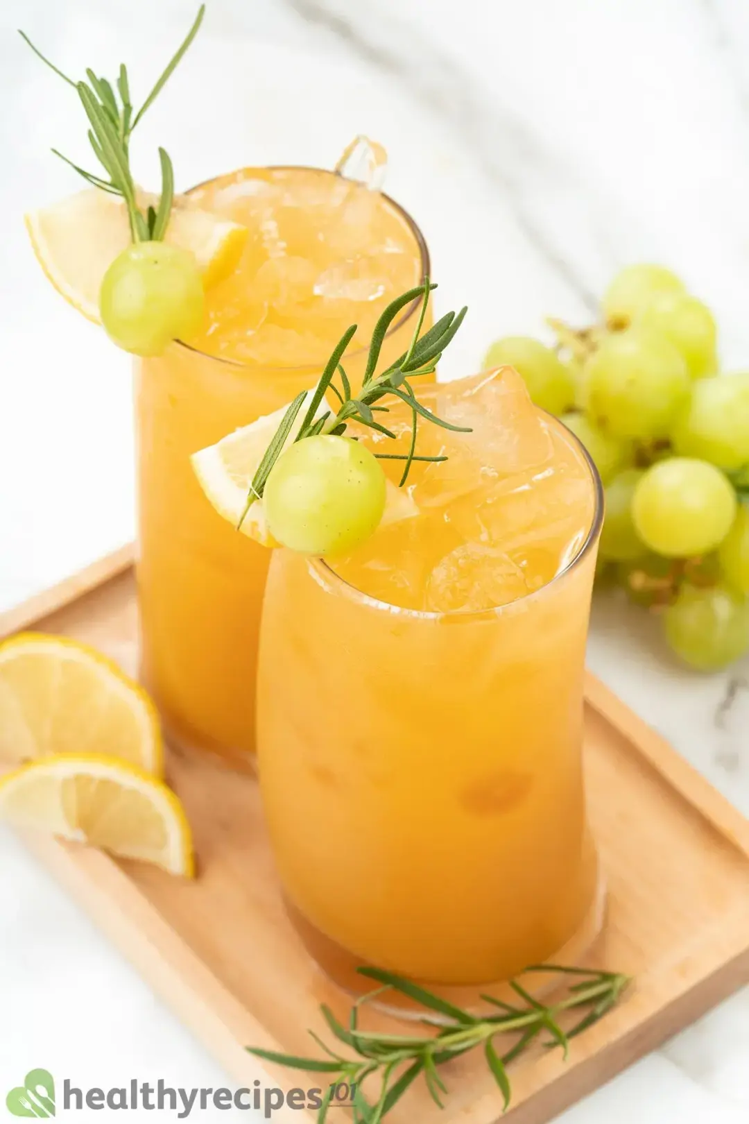 Two tall glasses of iced grapefruit drink, each topped with rosemary sprig, grape, lemon wedge, put on a wooden tray