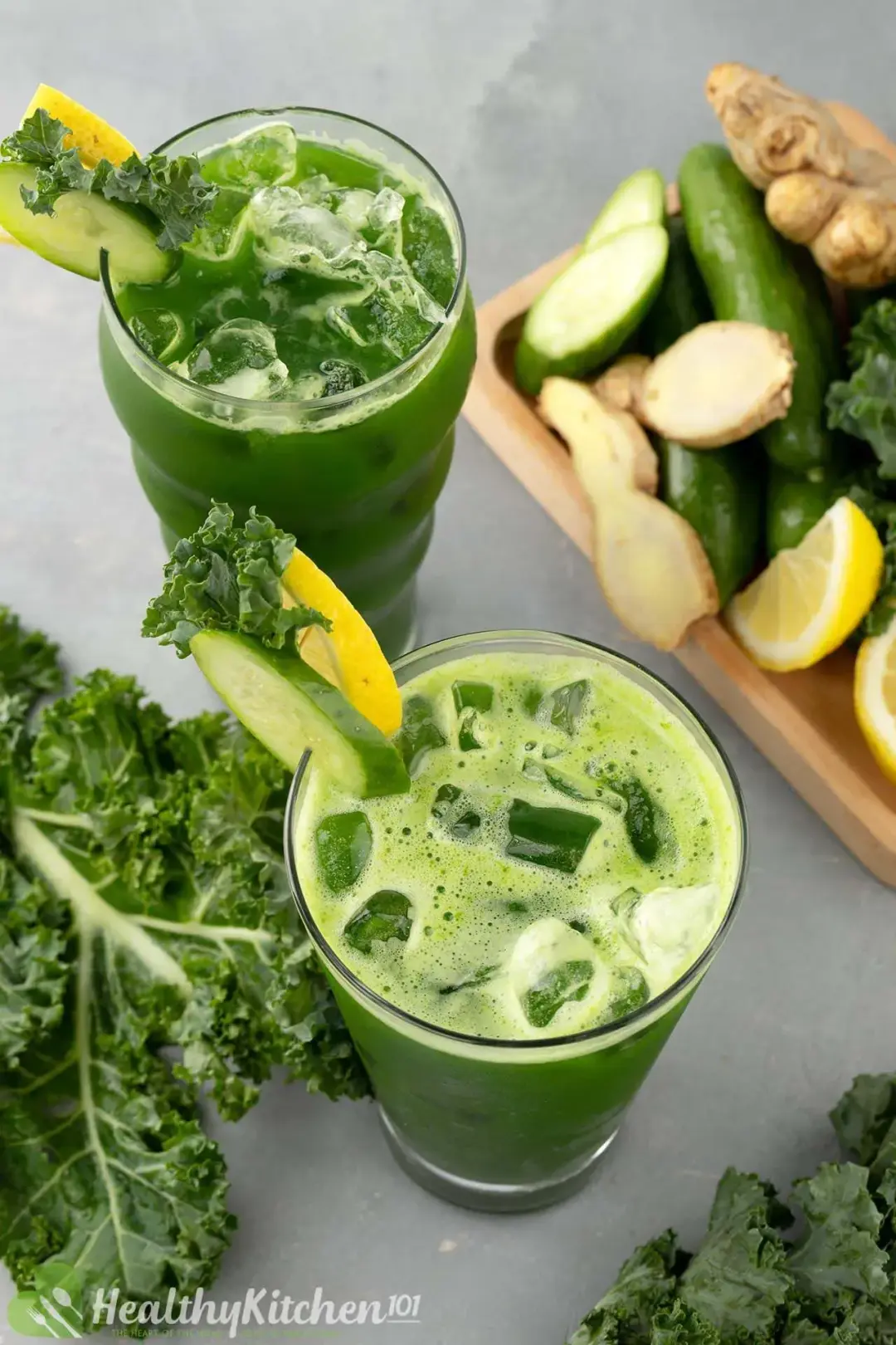 Two iced glasses of green kale juice next to some kale and a tray of cucumber, ginger, and lemon