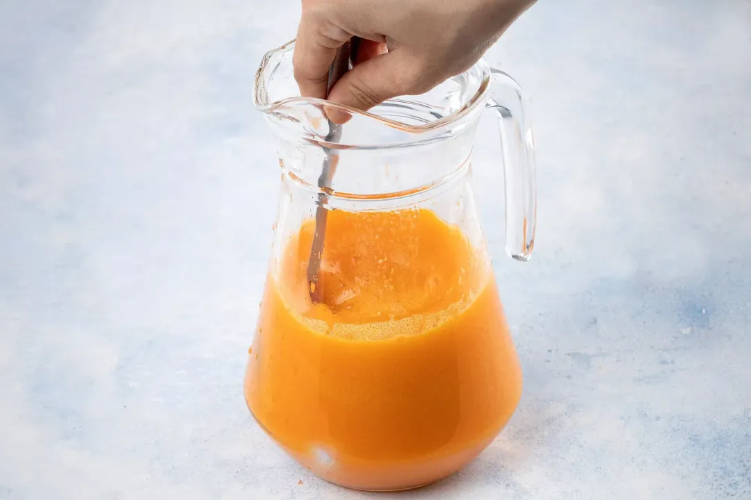 An orange drink mixed with a silver spoon in a glass pitcher