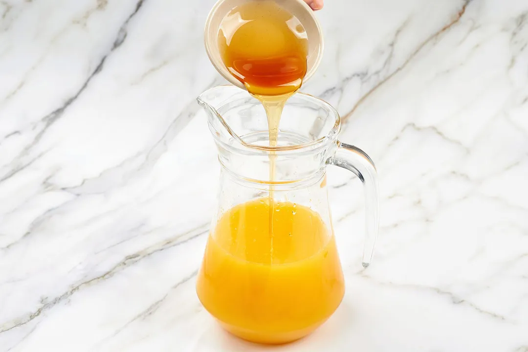 pouring honey from a small bowl into a glass pitcher of orange liquid
