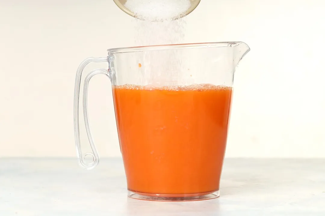 pouring sugar from a small bowl into a pitcher of orange liquid