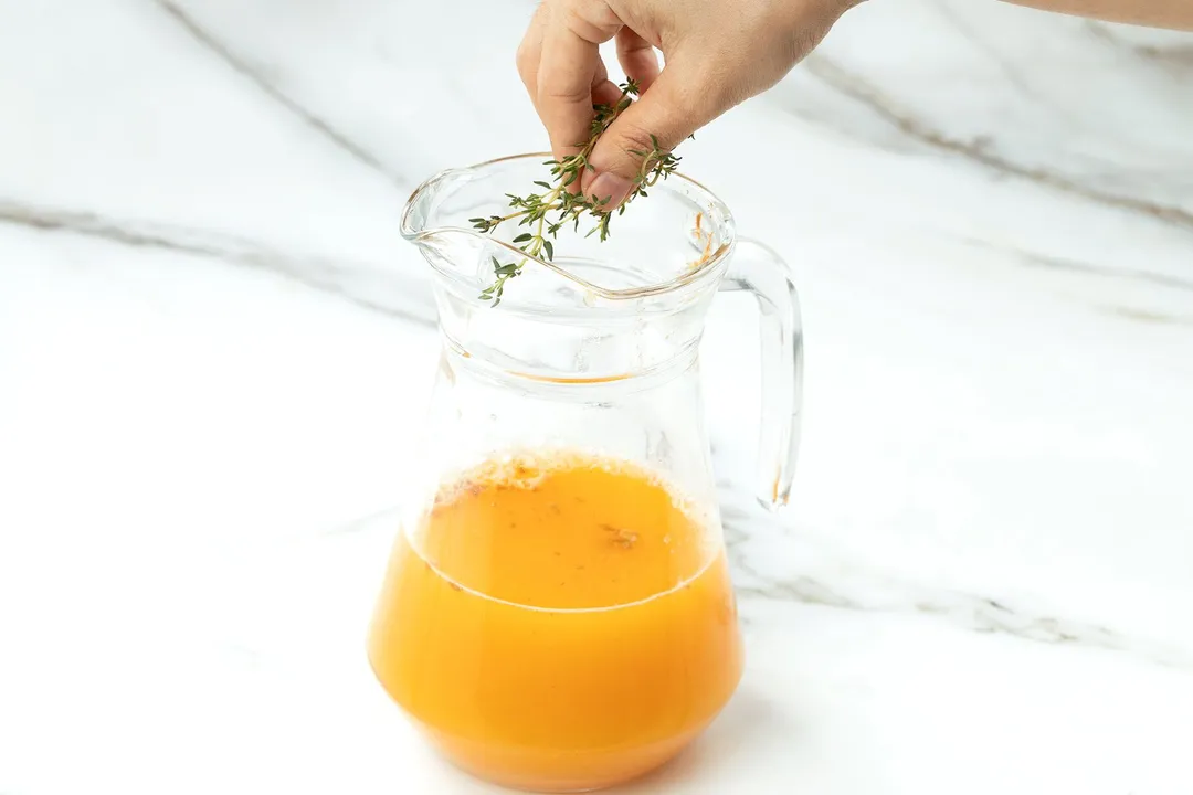 a hand holding thyme leaves on top of a pitcher of orange juice