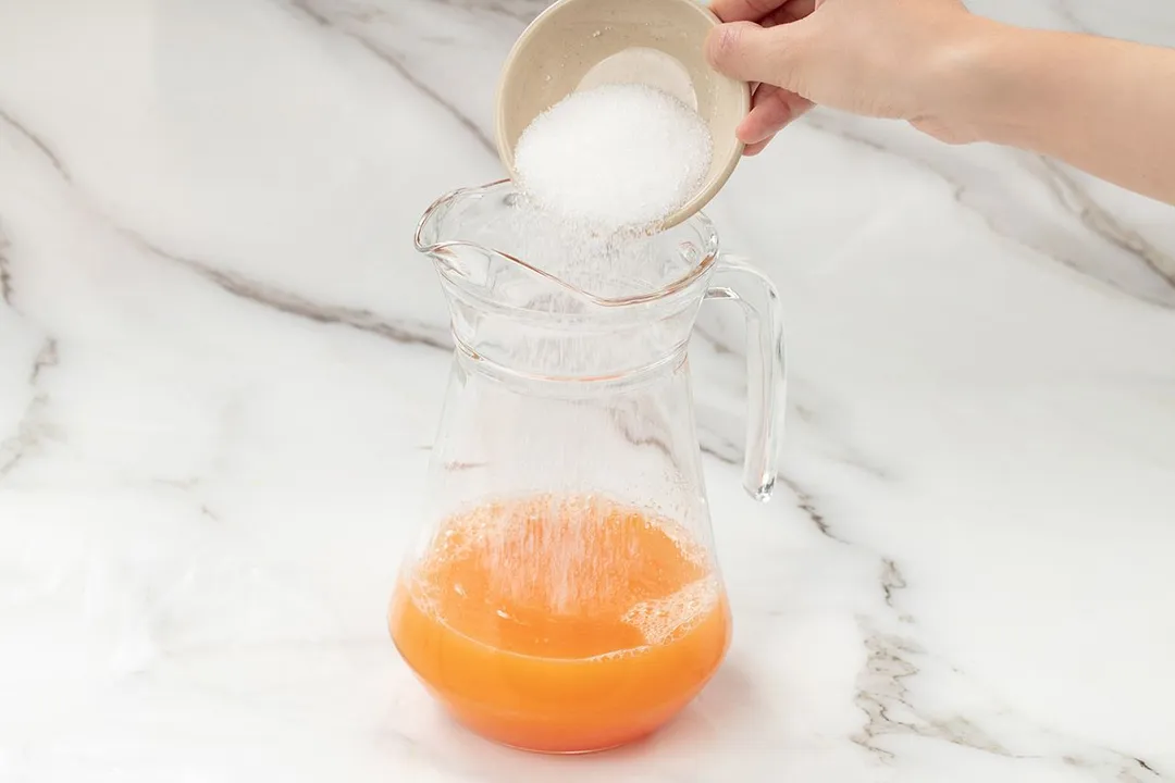 pouring sugar from a small bowl to a glass pitcher of juice