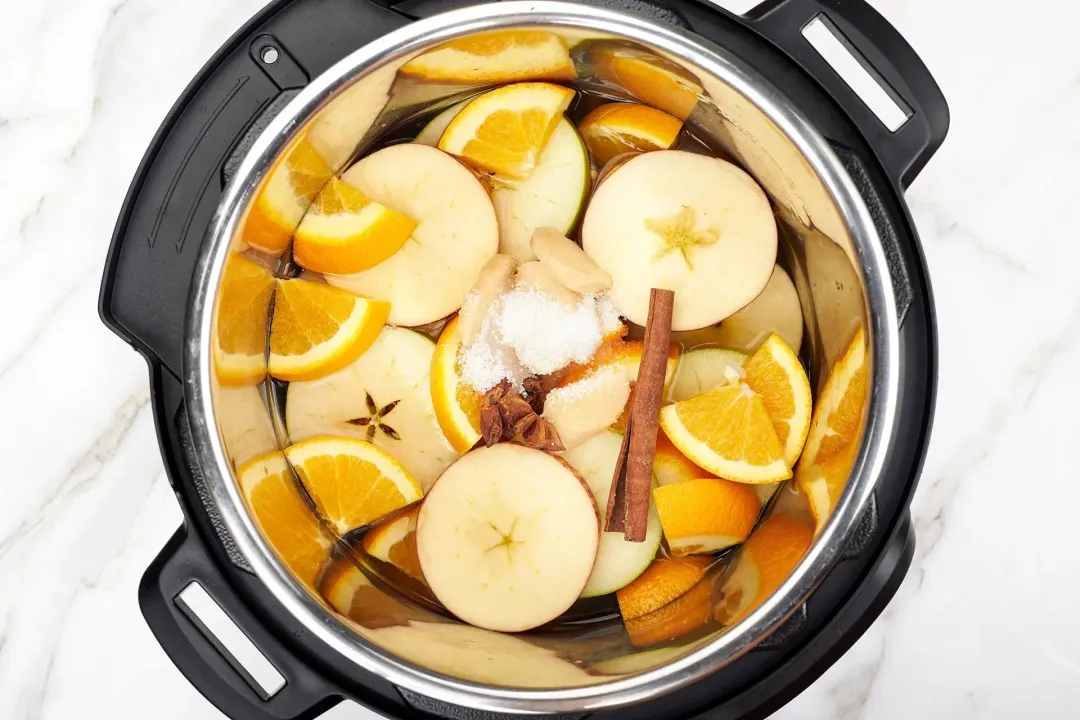In an instant pot: apple slices, orange triangles, cinnamon stick, and sugar in the middle