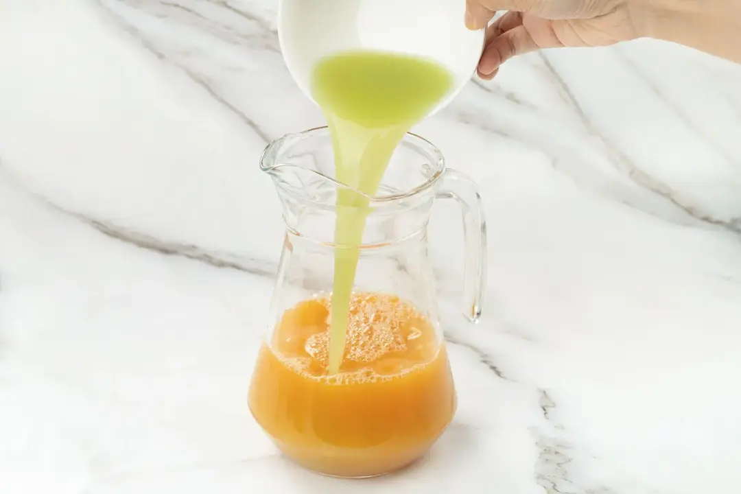 Pouring a green grape juice from a white bowl into a clear pitcher of grapefruit juice