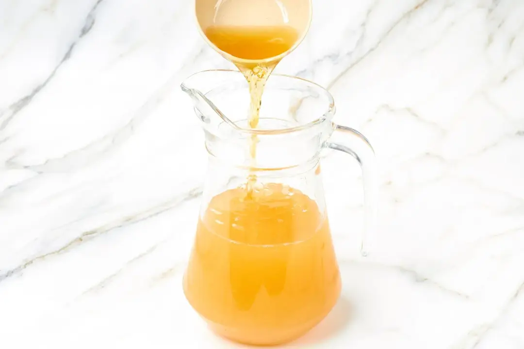A small bowl pouring whiskey from the top into a clear pitcher of apple cider cocktail