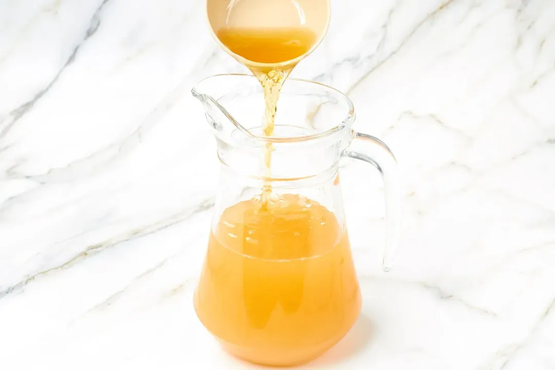 A small bowl pouring whiskey from the top into a clear pitcher of apple cider cocktail