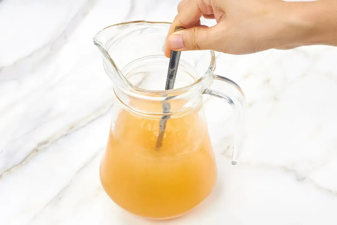 A glass pitcher of apple cider margarita stirred with a silver spoon by a hand