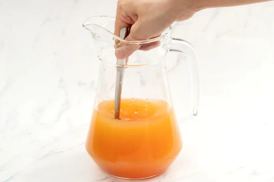 A clear pitcher of apple cider vinegar and grapefruit juice drink stirred with a silver spoon by a hand