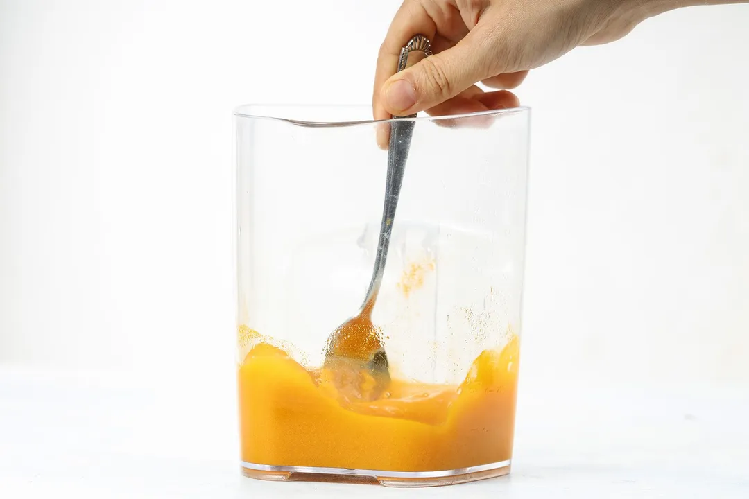 A hand using a spoon to stir Carrot Ginger Turmeric Juice in a large jug.