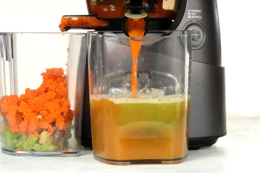 A juicer shooting out carrot juice into a pitcher of brown-green liquid, next to a container of carrot and cucumber pulps