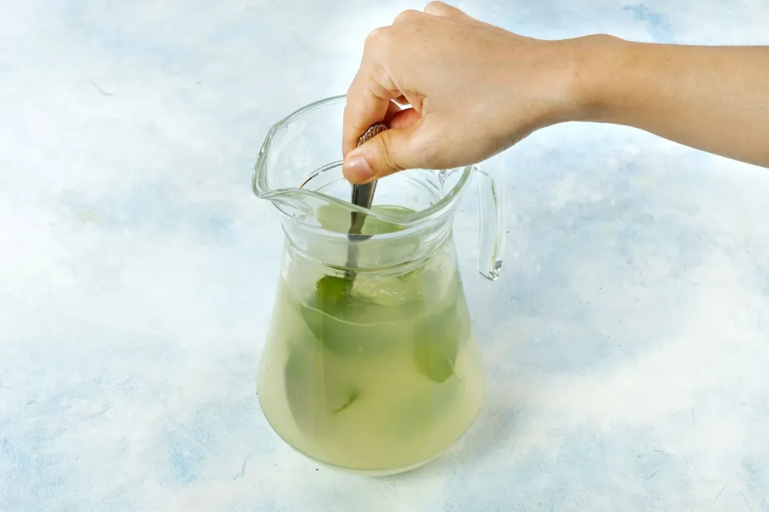 A spoon stirring a drink of lemon juice, ginger, and filled with mint leaves