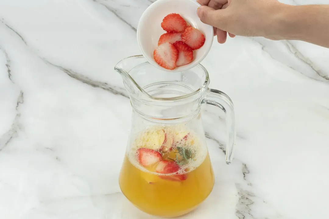 A pitcher filled with orange juice, fruits, and rosemary, with a bowl of sliced strawberries hovering on top