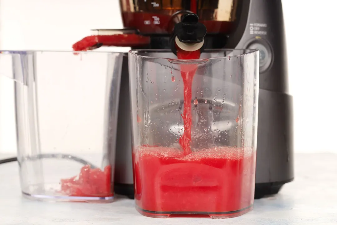 A juicer processing watermelon slices into juice. The liquid is poured into a large glass juice while the fruit remains are discarded into another.