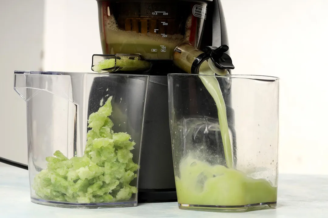 A juicer processing winter melon into a pale green juice in one glass jug and discarding the abundant materials into another.