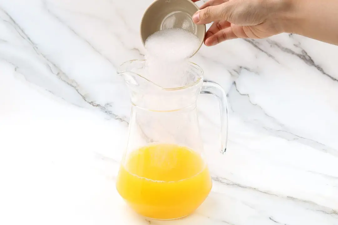 A bowl of sugar poured into a pitcher of orange juice and pineapple juice