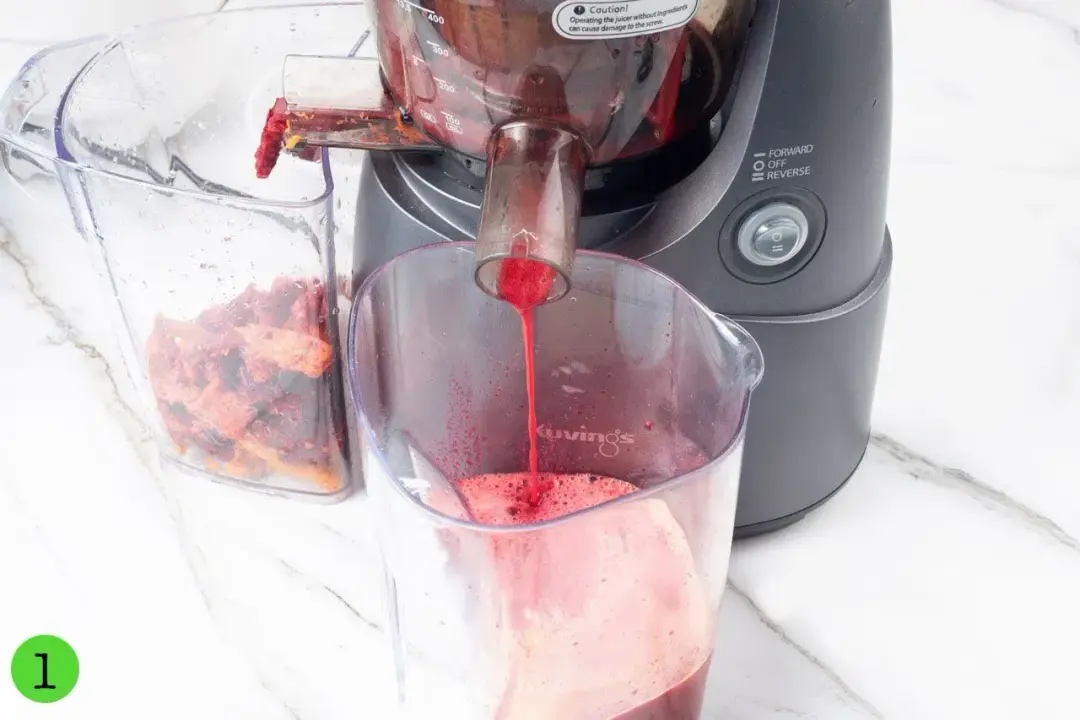 A juicer shooting out red juice into a clear pitcher on one end, and fruit pulp into a clear container on the other end