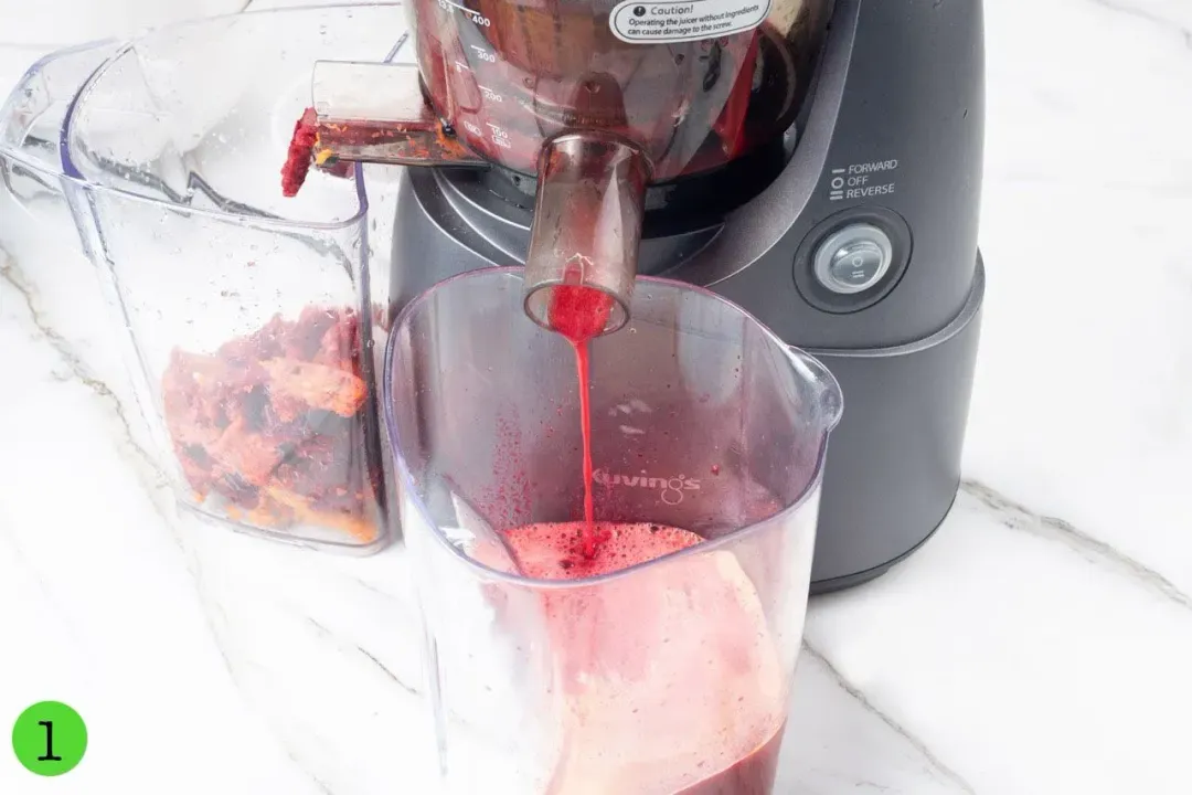 A juicer shooting out red juice into a clear pitcher on one end, and fruit pulp into a clear container on the other end