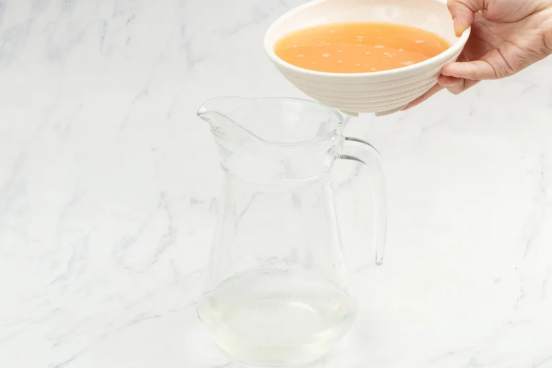 a hand holding a bowl of orange liquid on top of a glass pitcher