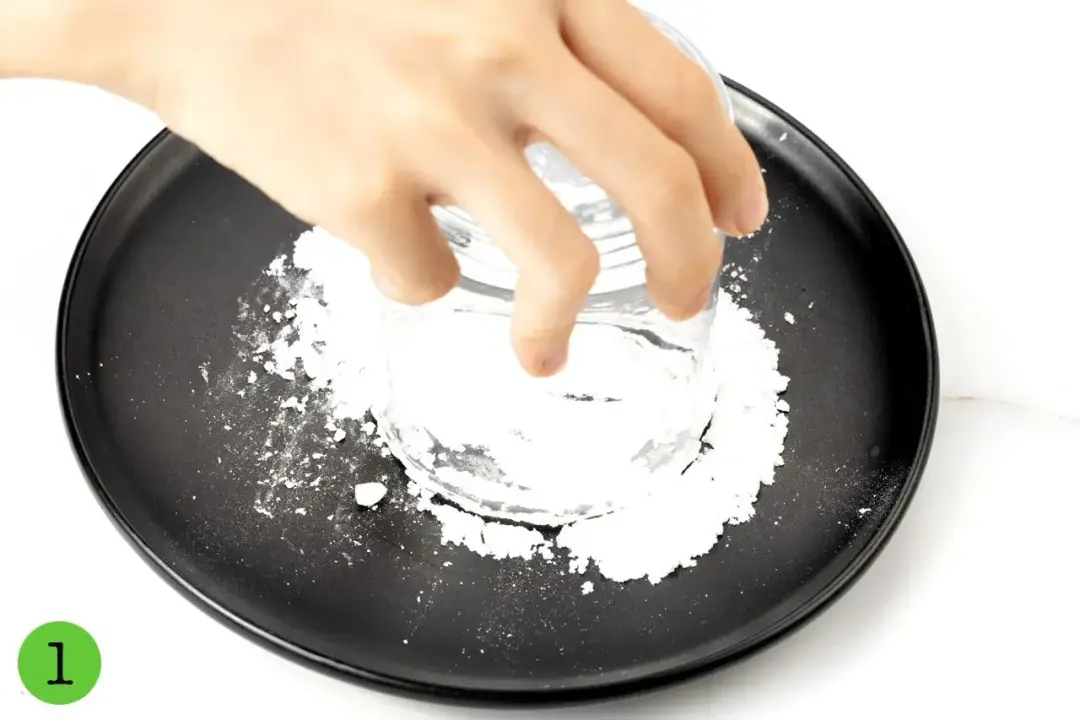 A hand dipping a short glass into powdered sugar on a black plate