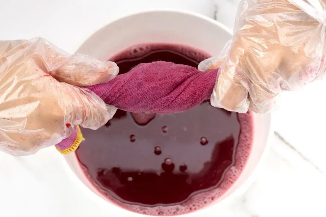 Two hands twisting a pomegranate-colored cheesecloth to extract pomegranate juice, collected in a white bowl