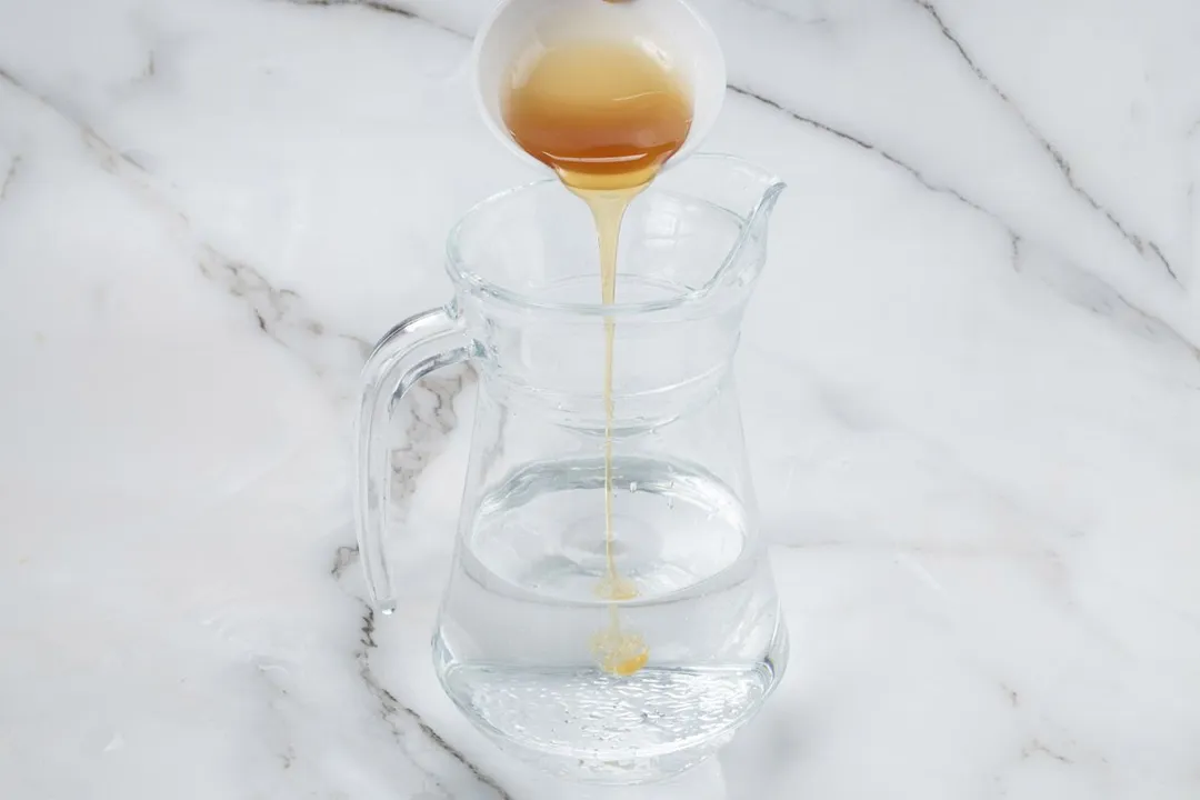 pouring honey from a small bowl into a glass pitcher of water