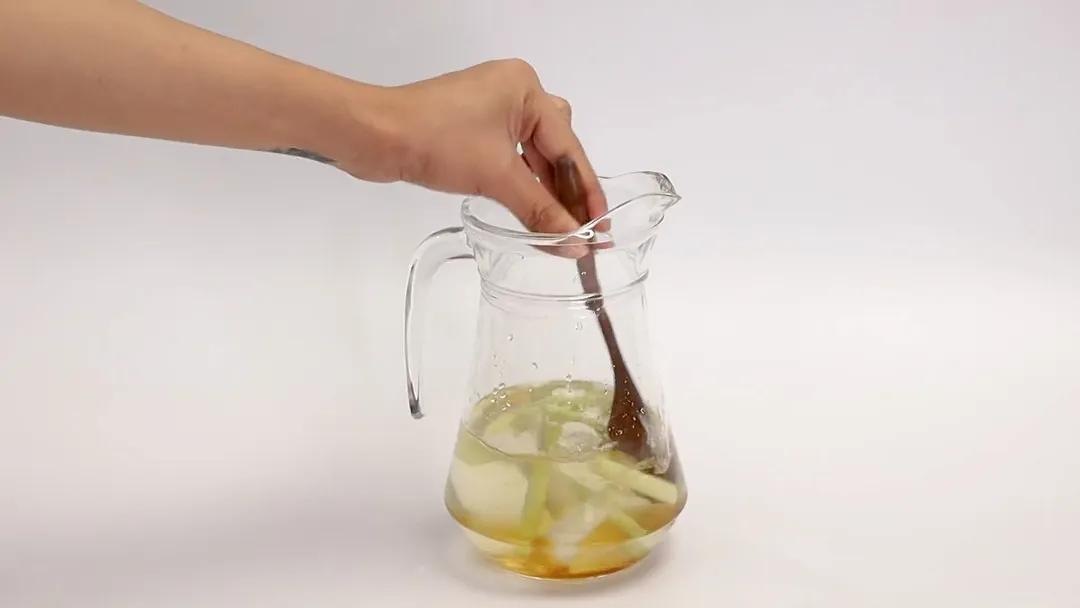 A hand holding a wooden spoon stirring mixture of apple cider vinegar and lemon juice with sliced lemongrass and honey in a glass jar.