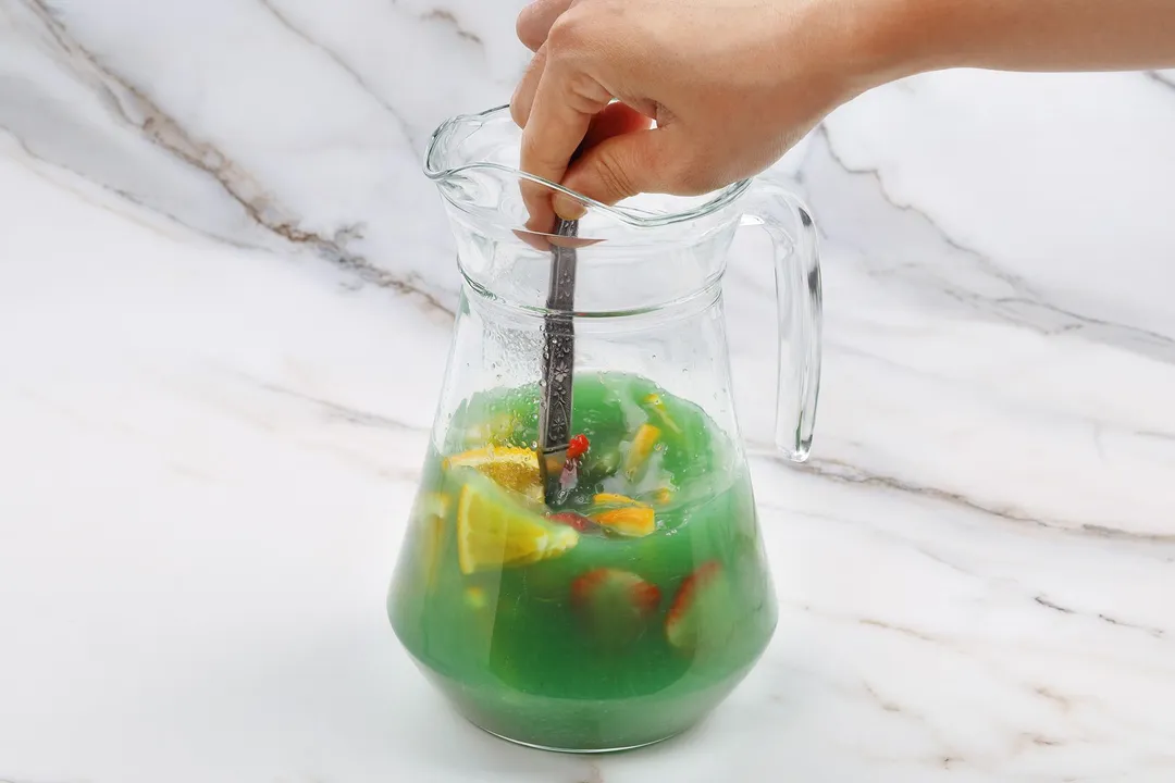 a hand holding a spoon stir a  large pitcher of green jungle juice