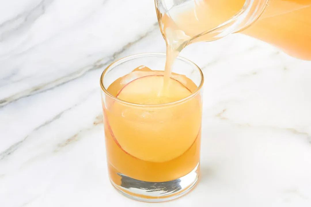 A pitcher pouring apple cider cocktail into a short glass filled with ice and apple slices