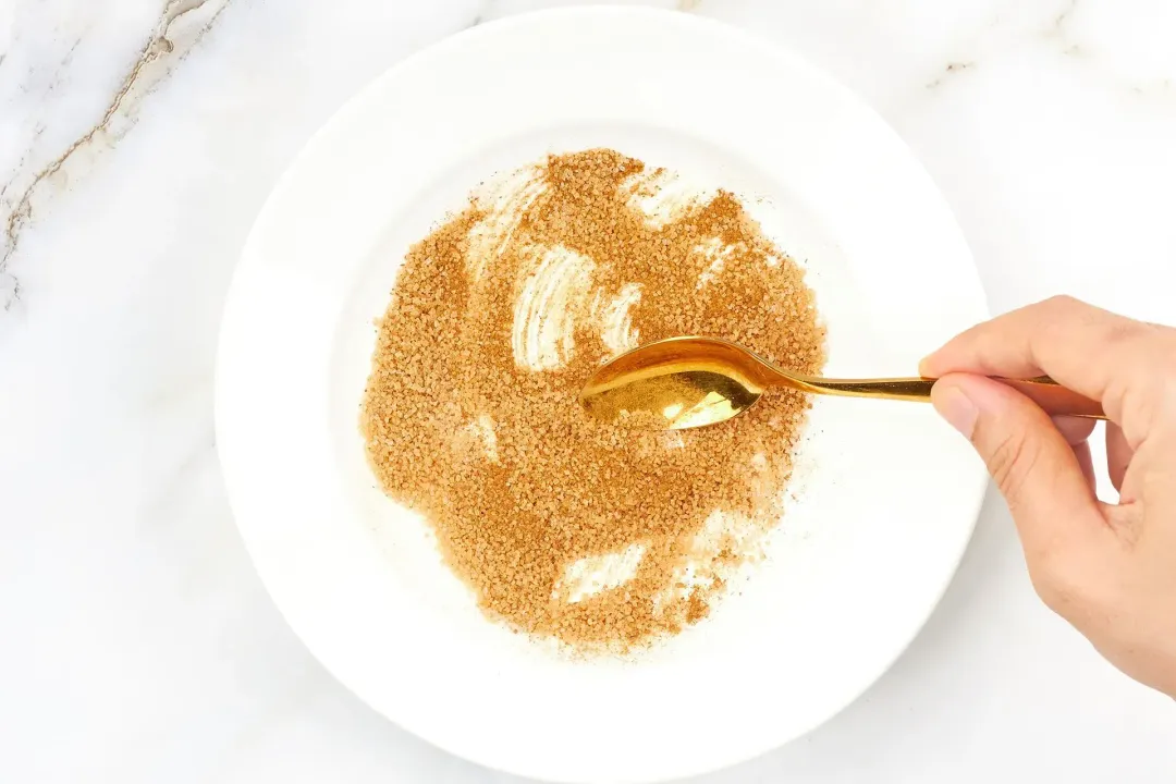 A brown sugar mixture in a white plate stirred with a golden spoon