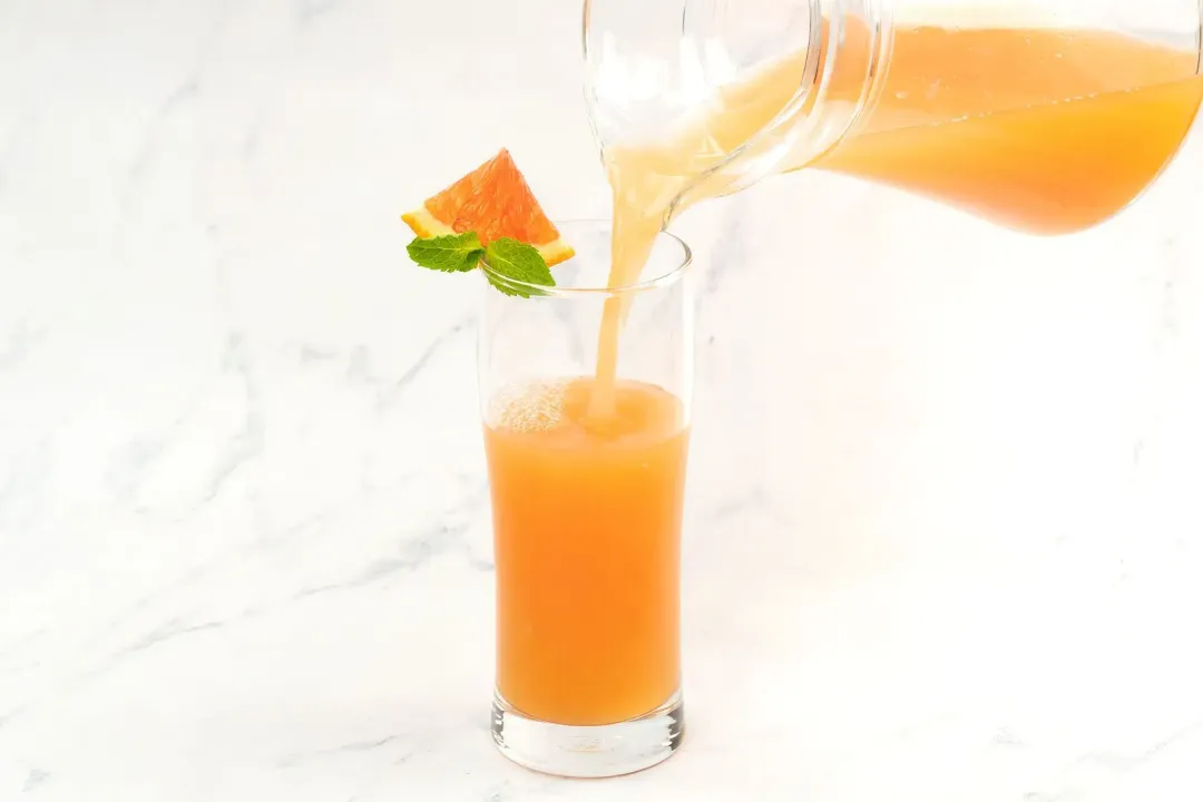 A clear pitcher pouring a cocktail of grapefruit juice and vinegar into a clear glass garnished with mints and a grapefruit wedge