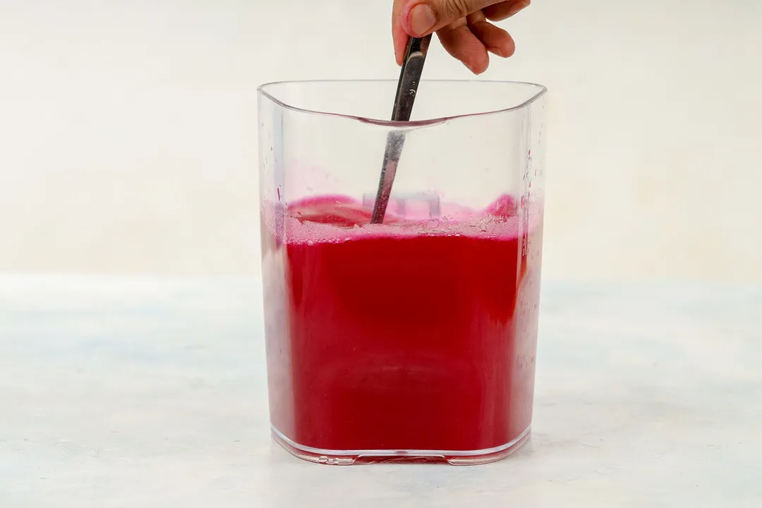 A hand using a spoon to stir in a large glass jug filled up to two-thirds with dragon fruit.